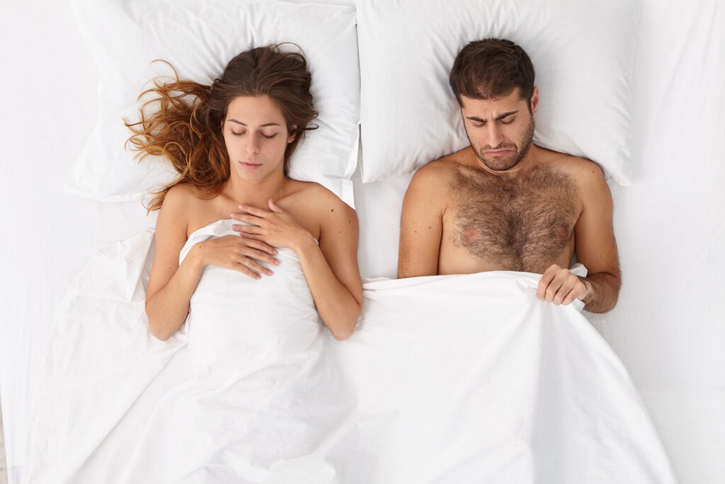 A woman lying next to a man in bed who is lifting the duvet because he has Erectile Dysfunction.